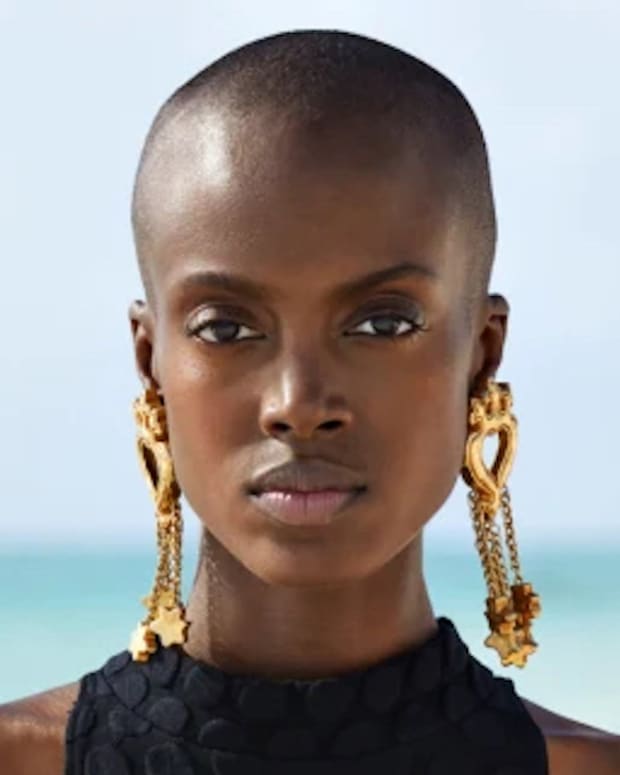 Madisin Rian poses in front of the ocean in a black onesie swimsuit and oversized gold earrings.